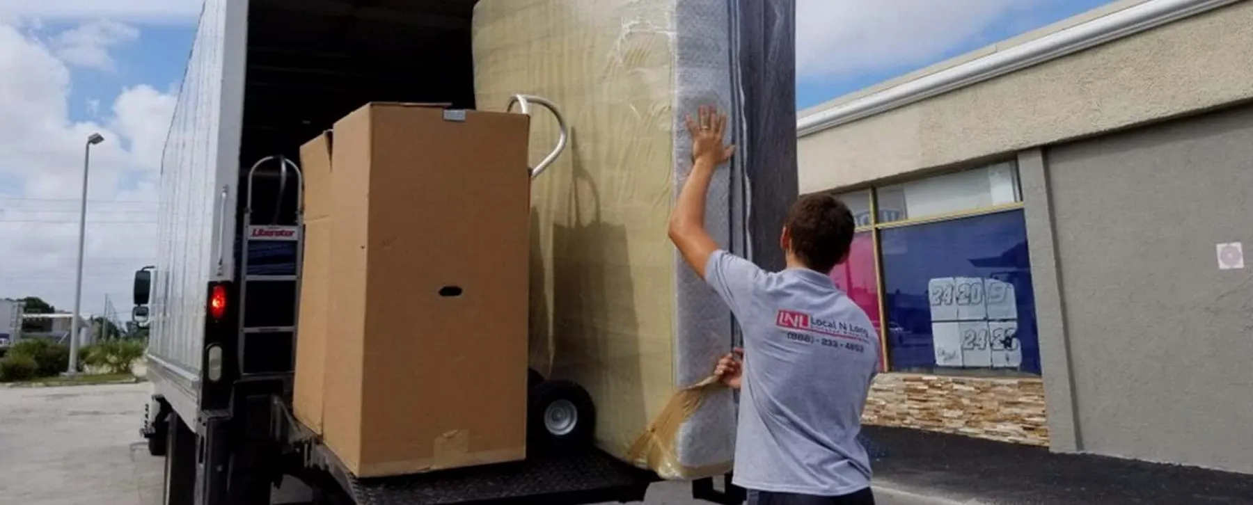Mover wearing an LNL company t-shirt loading a wrapped mattress onto a truck (wide crop)